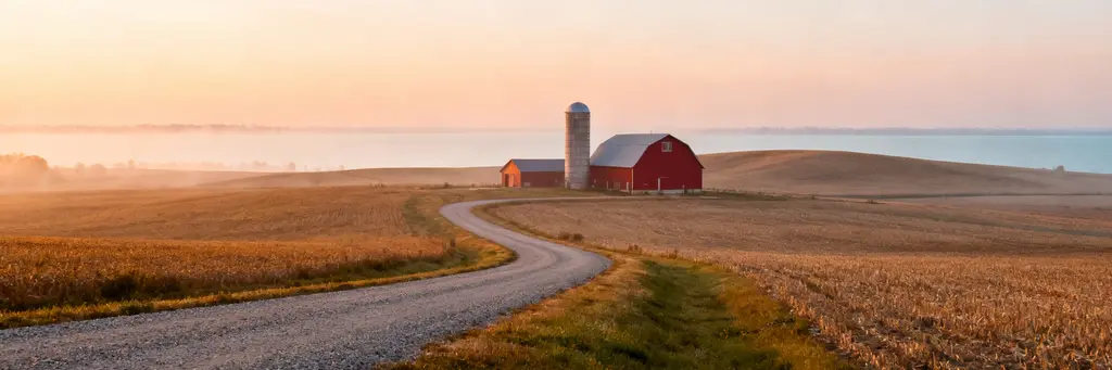 Rolling farmland with a red barn and silo along a gravel road at golden hour