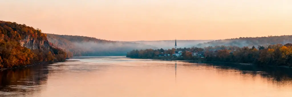 A calm river between wooded autumn bluffs under a peach sunrise sky