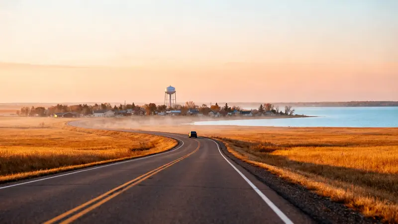A two-lane highway through golden prairie toward a small town with a water tower beside a lake